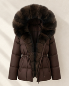 A dark brown puffer jacket with a large, luxurious dark brown fur-trimmed hood, shown against a neutral light beige background with soft shadow overlays.