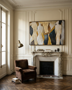 A large framed abstract painting with cream, gold, black, and gray shapes hangs above a white fireplace mantel in a classic room with cream paneled walls, a brown armchair, and a floor lamp.