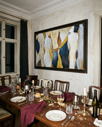 A large framed abstract painting with figures in cream, blue, and gold hangs on a distressed beige wall above a rustic wooden dining table set with plates, wine bottles, candles, and dark red napkins in a dimly lit dining room.