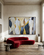 A large framed abstract painting featuring stylized human figures in cream, gold, navy, and beige tones, displayed on a textured beige wall above a curved deep red velvet sofa in a modern living room.
