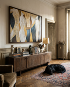 A large framed abstract painting with flowing cream, gold, navy, and gray shapes hangs on a beige wall above a wooden sideboard in a styled room. A black dog rests on a patterned rug on the floor below, with a lamp, books, and decor on the cabinet.