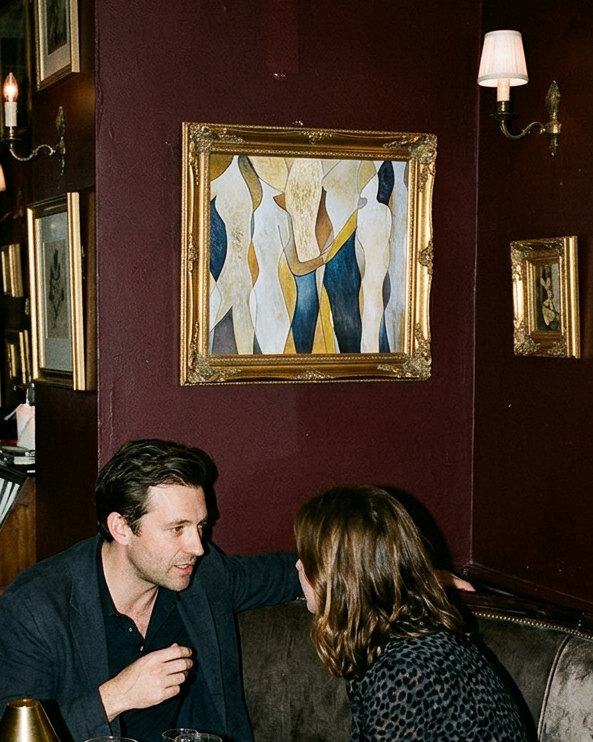 A man and a woman seated at a booth in a dimly lit restaurant or cafe, surrounded by framed wall art, candles, and tables with glasses and plates.