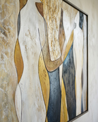 A framed abstract painting depicting stylized female figures in flowing cream, gold, brown, and slate blue-gray garments, shown hanging on a textured beige wall.