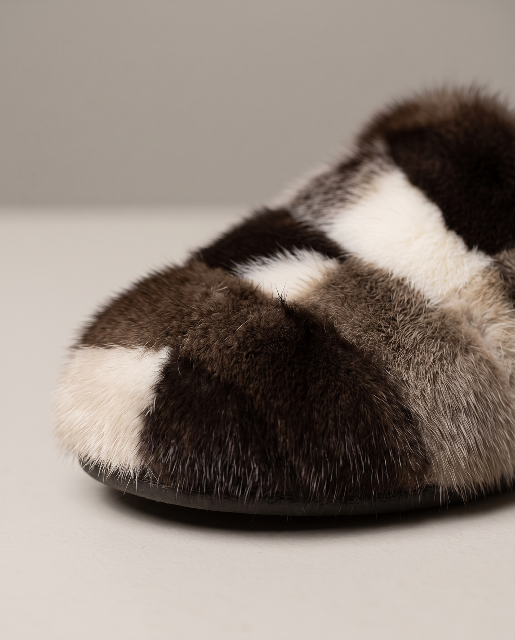 A close-up of a plush fur slipper with a patchwork pattern of dark brown, cream, and gray-brown, shown on a neutral light gray background.