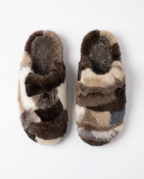 A pair of plush mule slippers made from patchwork animal fur, featuring a mix of brown, beige, cream, and gray tones, photographed from a top-down view on a light gray background.