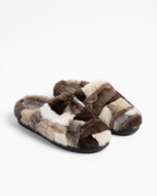 A pair of plush slip-on slippers made from patchwork faux fur in shades of brown, beige, cream, and white, with black soles, photographed on a clean white background.