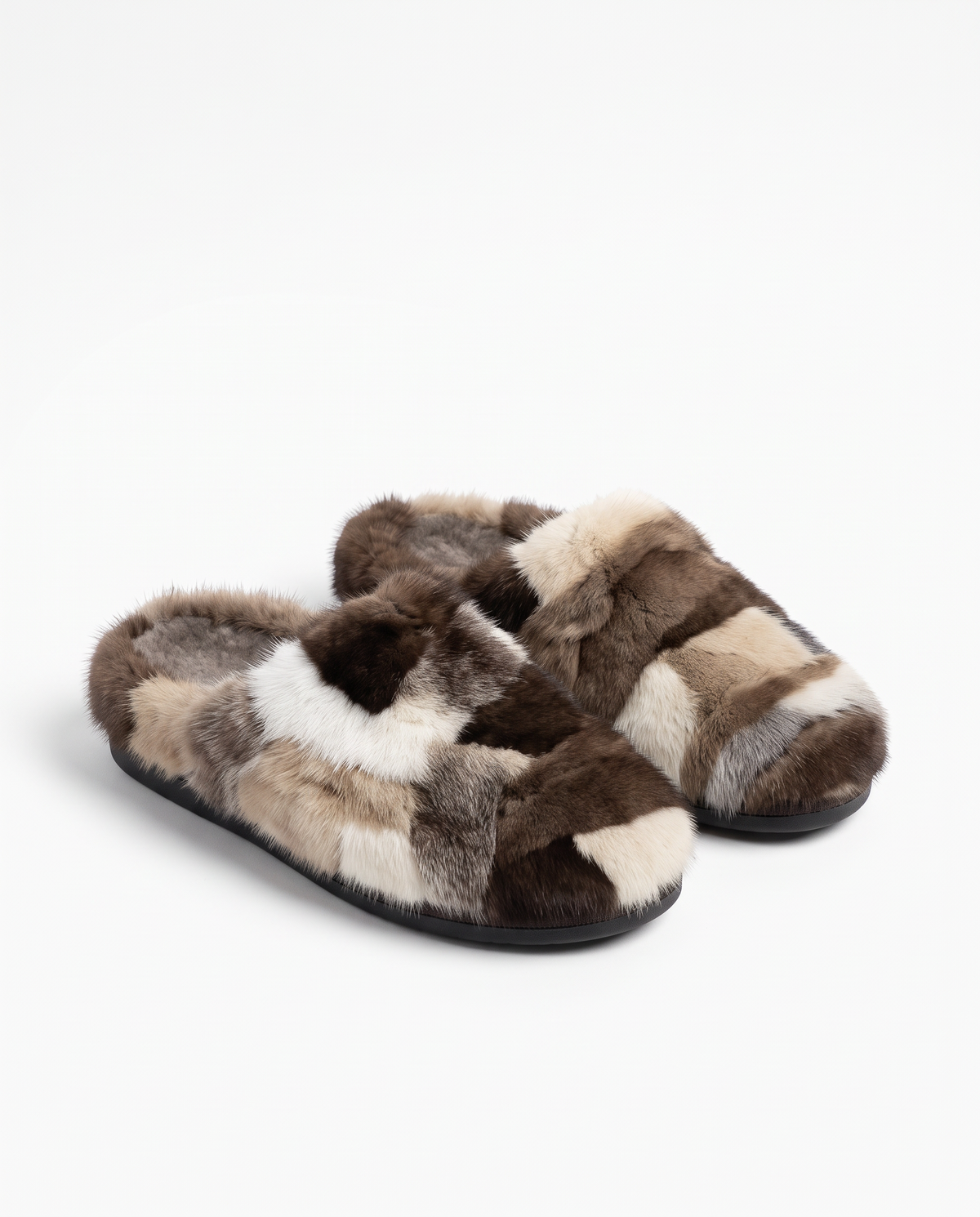 A pair of plush slip-on slippers made from patchwork faux fur in shades of brown, beige, cream, and white, with black soles, photographed on a clean white background.
