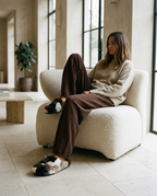 A woman sits in a cream boucle lounge chair in a bright, modern room, wearing a beige knit sweater, brown wide-leg trousers, and fluffy brown-and-white slippers.