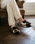 A person seated in a brown armchair wearing plush patchwork slippers in brown, cream, black, and white, standing on a dark wood herringbone floor.