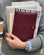 A person in a gray knit sweater holds a stack of books and magazines, with a burgundy ostrich-textured notebook cover in the center. The person also holds a gold pen and a takeaway coffee cup.