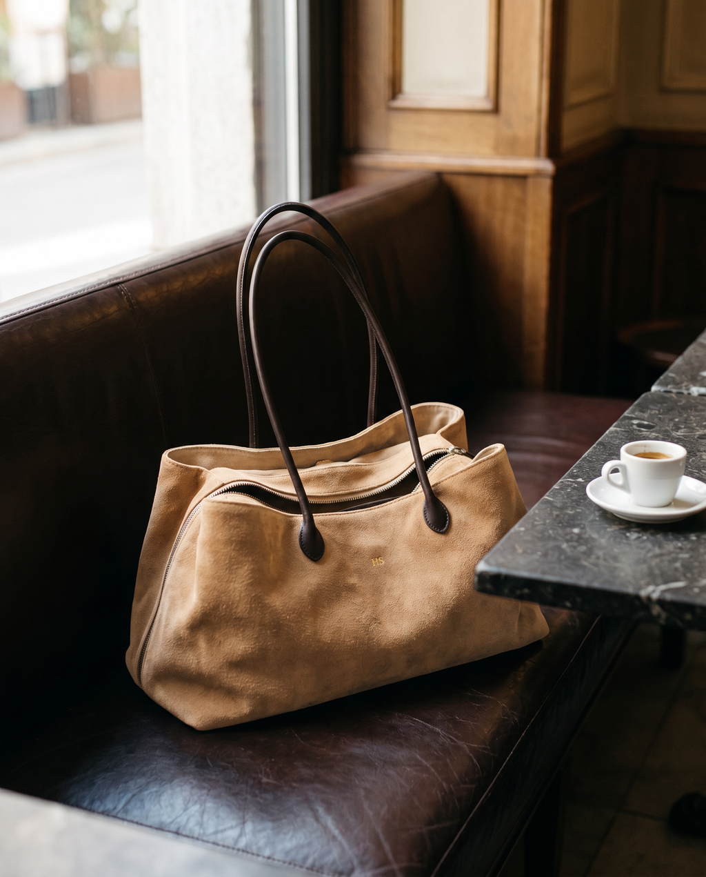 The Marais Tote in Apricot suede on a leather cafe banquette beside an espresso
