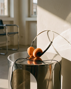 Orange over-ear headphones with black earcups and headband rests on a reflective silver metal side table in a sunlit room.