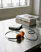 A pair of over-ear headphones with bright orange ear cushions and black cables lies on a brushed metal table. A white handheld cassette player, a stack of magazines, and a glass of water are nearby, with a softly lit window in the background.