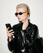 A blonde model wearing a black leather jacket, black sunglasses, and orange over-ear headphones, holding a black smartphone against a plain off-white background.