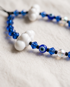 A close-up of a beaded necklace featuring blue glass beads, white cloud-shaped beads, a blue evil eye bead, and clear faceted beads, resting on a textured beige fabric background.