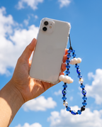 A hand holds a clear smartphone case with a blue beaded phone strap featuring evil eye beads and white cloud charms against a bright blue sky with clouds.