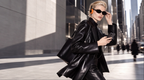 A woman walking down a city street wearing a shiny black leather jacket and matching black leather pants, black sunglasses, and orange over-ear headphones, carrying a black shoulder bag.