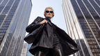 A woman in a black leather blazer and matching pants walking between tall glass skyscrapers, wearing orange over-ear headphones and black sunglasses.