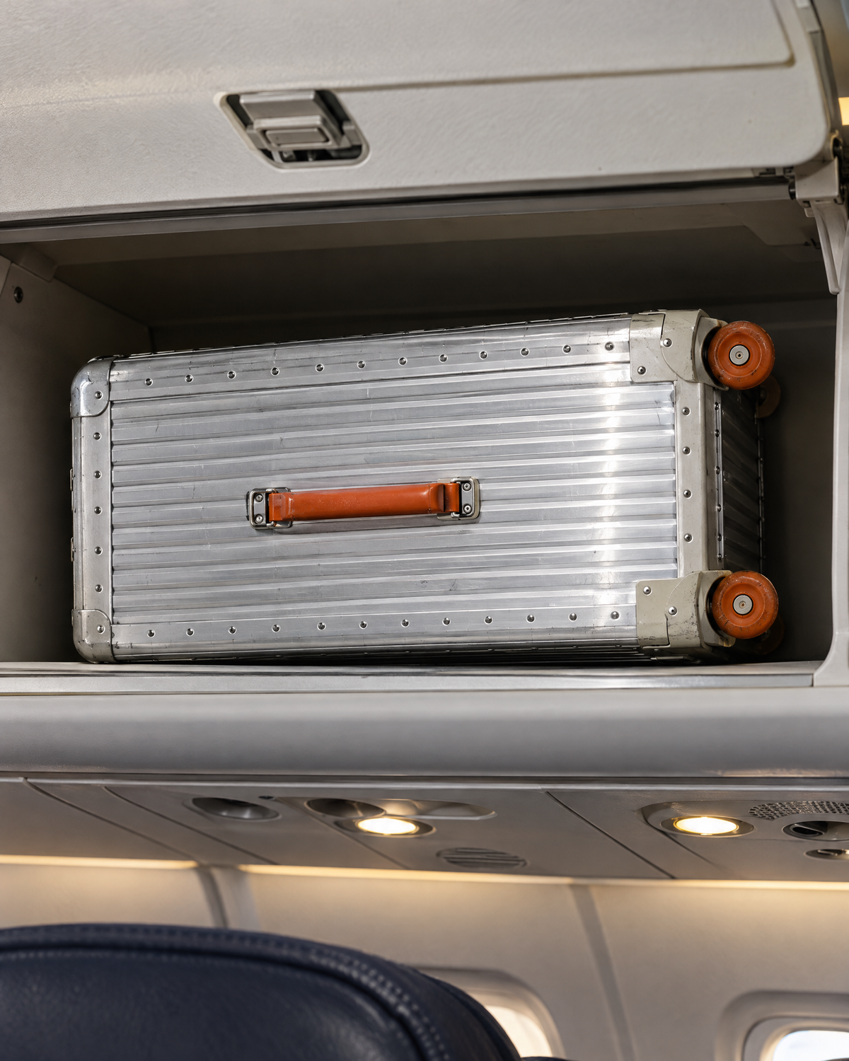 The Meridian Suitcase in silver with orange in an overhead compartment