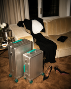 Two silver hard-shell suitcases with turquoise handles and wheels are placed on a brown carpet in a living room setting, with a beige sofa, black clothing, and black heels nearby.