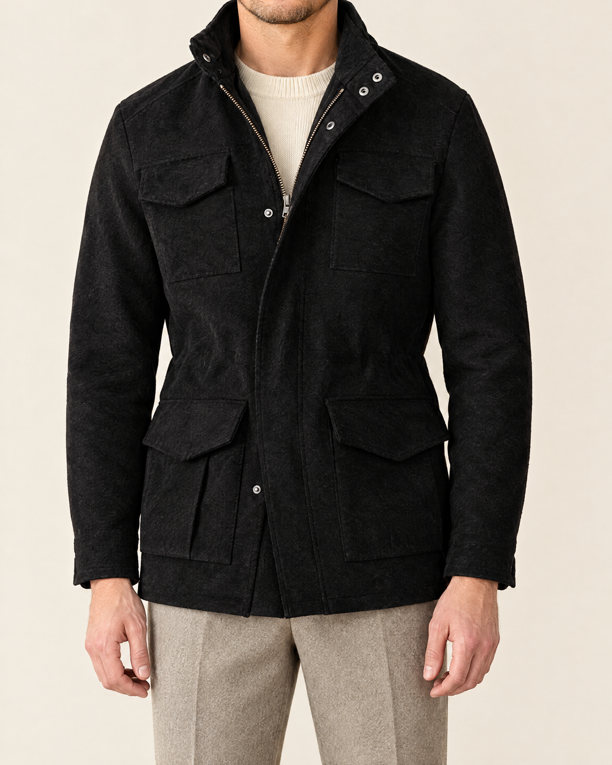 A man wearing a black textured wool-blend jacket with a stand collar, front zipper, and four flap pockets, styled over a cream knit sweater and light gray trousers, against a solid beige background.