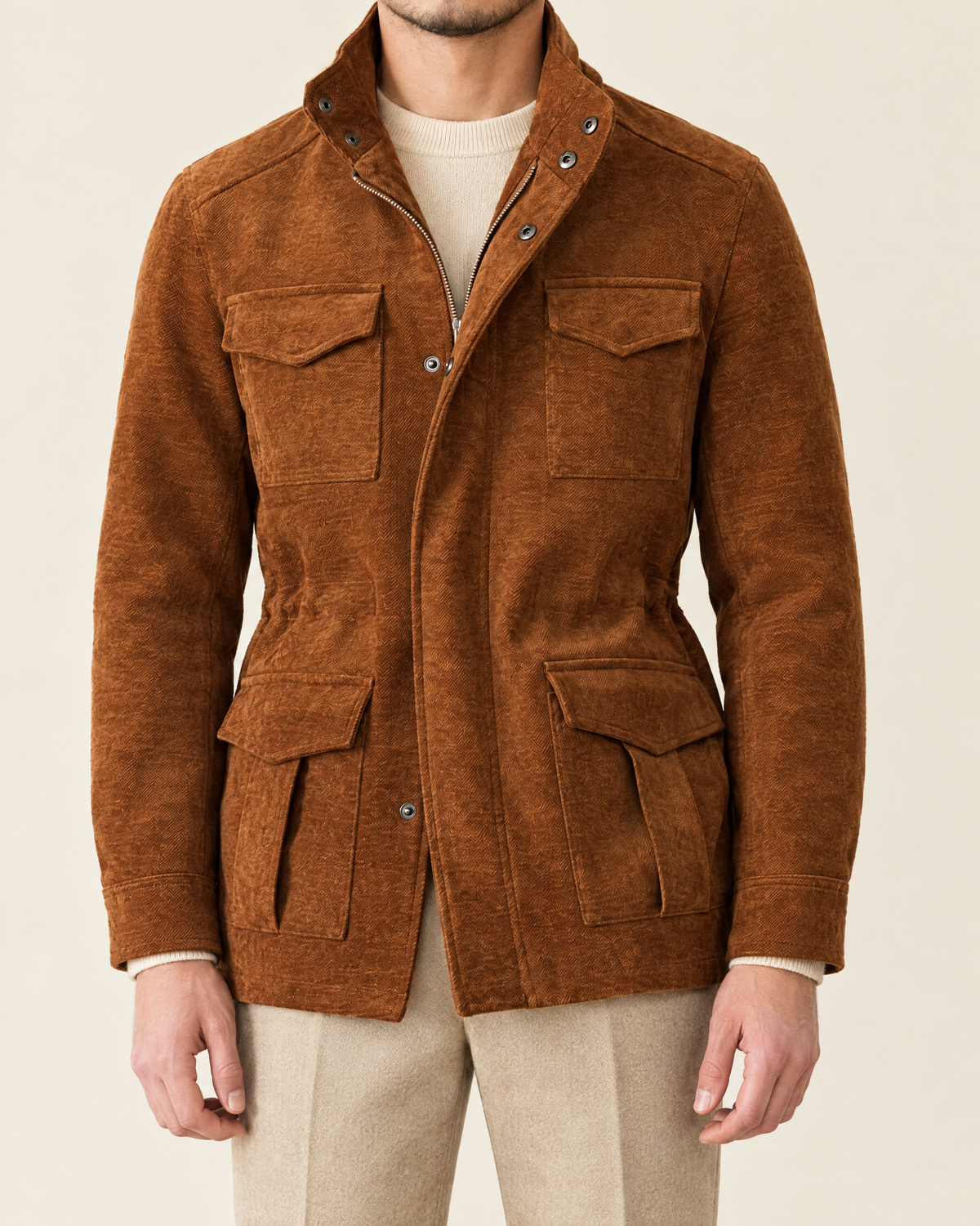A man wearing a brown corduroy jacket with a stand-up collar, snap-button front, and four front flap pockets, paired with a cream sweater and beige trousers, against a light beige background.