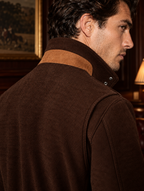 A man shown from the back and side wearing a dark brown corduroy jacket with a contrasting tan suede-like collar, set in a warm, dimly lit indoor environment.