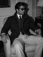 A man wearing a dark jacket, striped tie, and light trousers sits in an armchair holding a glass, photographed in a dimly lit indoor setting.