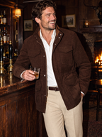 A man in a brown corduroy jacket, white shirt, and beige trousers stands in a dimly lit bar, holding a glass of whiskey.
