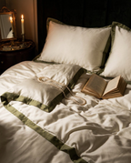 A styled bed with cream bedding and olive green satin trim, set in a dimly lit bedroom with a lit candle, an open book, and a pearl necklace on the duvet.
