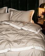 A beige bedding set with white piping trim, styled on a bed in a cozy bedroom setting with a dark green velvet headboard, a lit gold lamp, and a glass of red wine on a wooden nightstand.