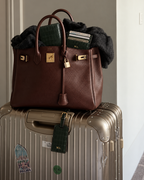 A brown pebbled leather handbag with gold hardware sits on top of a metallic suitcase, filled with a dark green crocodile-embossed wallet, cards, and a dark fur accessory. The handbag features a front zip closure, a hanging leather strap, and a padlock, with an 'ML' luggage tag attached to the suitcase.