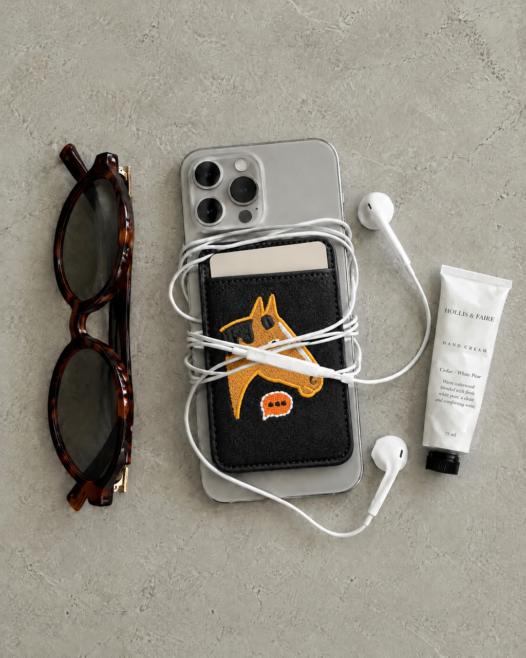 The Gallop Card Holder in horse patch black on the back of a phone, holding a card, with earphones, sunglasses and hand cream