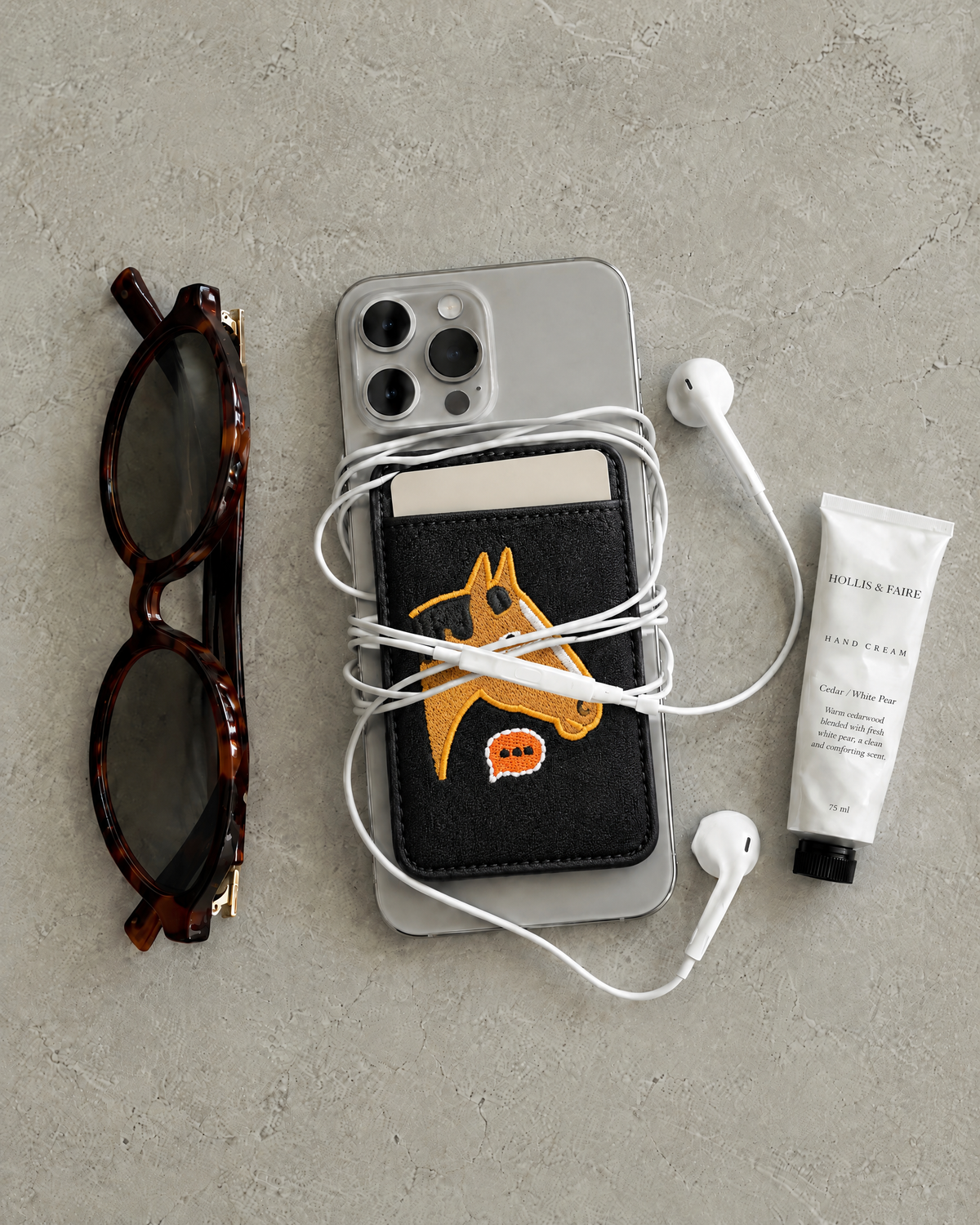 The Gallop Card Holder in horse patch black on the back of a phone, holding a card, with earphones, sunglasses and hand cream