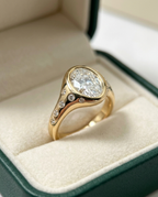 A yellow gold ring with a large oval-cut clear center stone and small accent stones along the shank, displayed in an open dark green jewelry box with cream velvet lining.