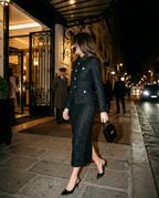 A woman walks down a city street at night wearing a dark tweed two-piece suit with silver-tone buttons, black heels, and carrying a small black handbag.