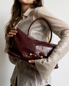 A woman in a shimmering beige ribbed blouse holds a burgundy crocodile-embossed leather handbag with gold-tone hardware against a plain light background.