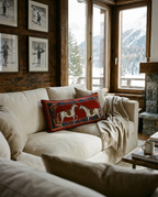 A cozy living room scene with a cream sofa, a red decorative throw pillow featuring two white horses, a draped cream blanket, and large windows showing a mountain view.