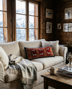 A cozy living room scene with a cream upholstered sofa, a red patterned throw pillow featuring white horses, a gray knit throw blanket, and a rustic wooden coffee table in front. Large wood-framed windows reveal a snowy mountain landscape outside, and framed wall art hangs on the rustic wood-paneled wall.