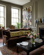 A styled living room featuring a dark brown velvet loveseat with two green patterned accent pillows, set in front of a black coffee table with a pink floral arrangement. The room has classic paneled walls, a fireplace, and a large abstract painting.