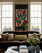 A styled living room featuring a dark brown velvet sofa with patterned throw pillows, set against a dark wall with a colorful abstract painting. Two tall windows with white frames are visible behind the sofa, and a black coffee table with books and a vase of flowers sits in the foreground on a patterned rug.