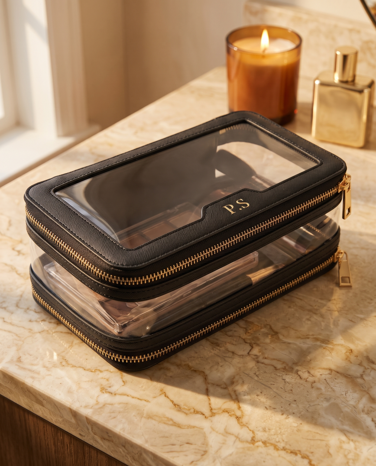 A black zippered makeup organizer case with a clear top window and gold-tone hardware, placed on a marble countertop beside a lit candle and a perfume bottle.