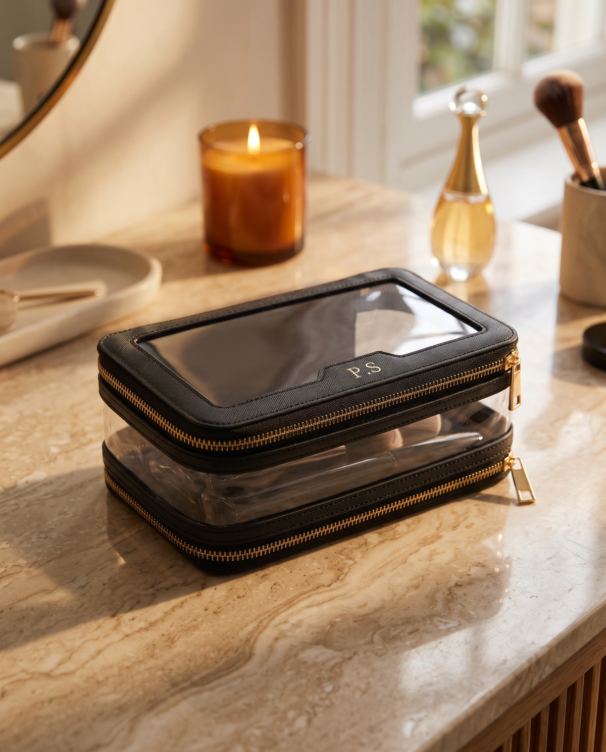 A black and clear zippered travel organizer case with gold-tone zippers, shown stacked on a marble vanity surface in a warm, softly lit room with a candle, makeup brushes, and a small bottle in the background.