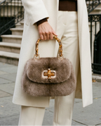 A model wearing a long cream coat, brown sweater, and cream trousers carries a brown fur handbag with gold-tone hardware in an urban street setting.