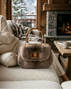 A woman in a cozy wooden cabin lounge setting, wearing a cream knit sweater and sitting on a textured beige sofa next to a brown fur handbag with a bamboo handle and gold hardware.