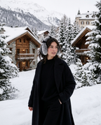 A woman wearing a long black coat and gray fuzzy earmuffs stands in a snowy mountain village with wooden chalets and snow-covered trees in the background.