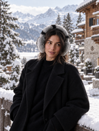 A woman wearing a black coat and gray-and-white patterned earmuffs stands outdoors in a snowy mountain setting with a chalet and trees in the background.