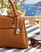A brown leather handbag with a silver padlock charm sits on a blue-and-white striped tablecloth outdoors. A wine glass, tortoiseshell sunglasses, and a scenic sea view are visible in the background.