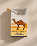 A white ceramic cigarette ashtray featuring a vintage-style illustration of a camel in a desert with the text 'TURKISH & AMERICAN BLEND CIGARETTES.' A lit cigarette rests in one of the ashtray slots.