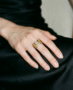 A close-up of a hand wearing a gold ring with a central cluster of clear gemstones, resting on a black satin fabric surface.